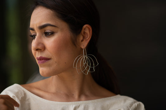 A model with long dark silky hair shows off a pair of statement earrings by American-Belgian jewelry designer Hannah Tomoko. These earrings are large but light and they close in a nearly invisible tubing to enhance the continuity of the design. Each earring is made of one continuous sterling silver wire that is meticulously curved and bent. This particular model is like a a wave of curved flower petals or fanning bird feathers.