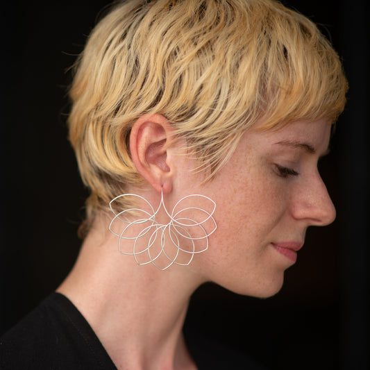 A pale model with cute short blond hair stands in front of a black wall of the US Federal courthouse in Austin, Texas. The eyecatcher: a pair of silver statement earrings by American-Belgian jewelry designer Hannah Tomoko. One of these earrings fills up the palm of a hand, but is surprisingly lightweight. The design is like a floral fan, or fanning bird tail, like a wave of feathers or petals flaring along the model's chin.