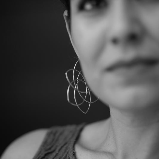 A black and white image of a model showing off a pair of flower earrings by American-Belgian jewelry designer Hannah Tomoko. Like a geometric 6-petal flower, these earrings are balanced not only visually but also when they are worn: they are large but light and they close in a nearly invisible tubing to enhance the continuity of the design. Each earring is made of one continuous sterling silver wire that is meticulously curved and bent. The flower is visible from the side, and is more like a hoop frontally.
