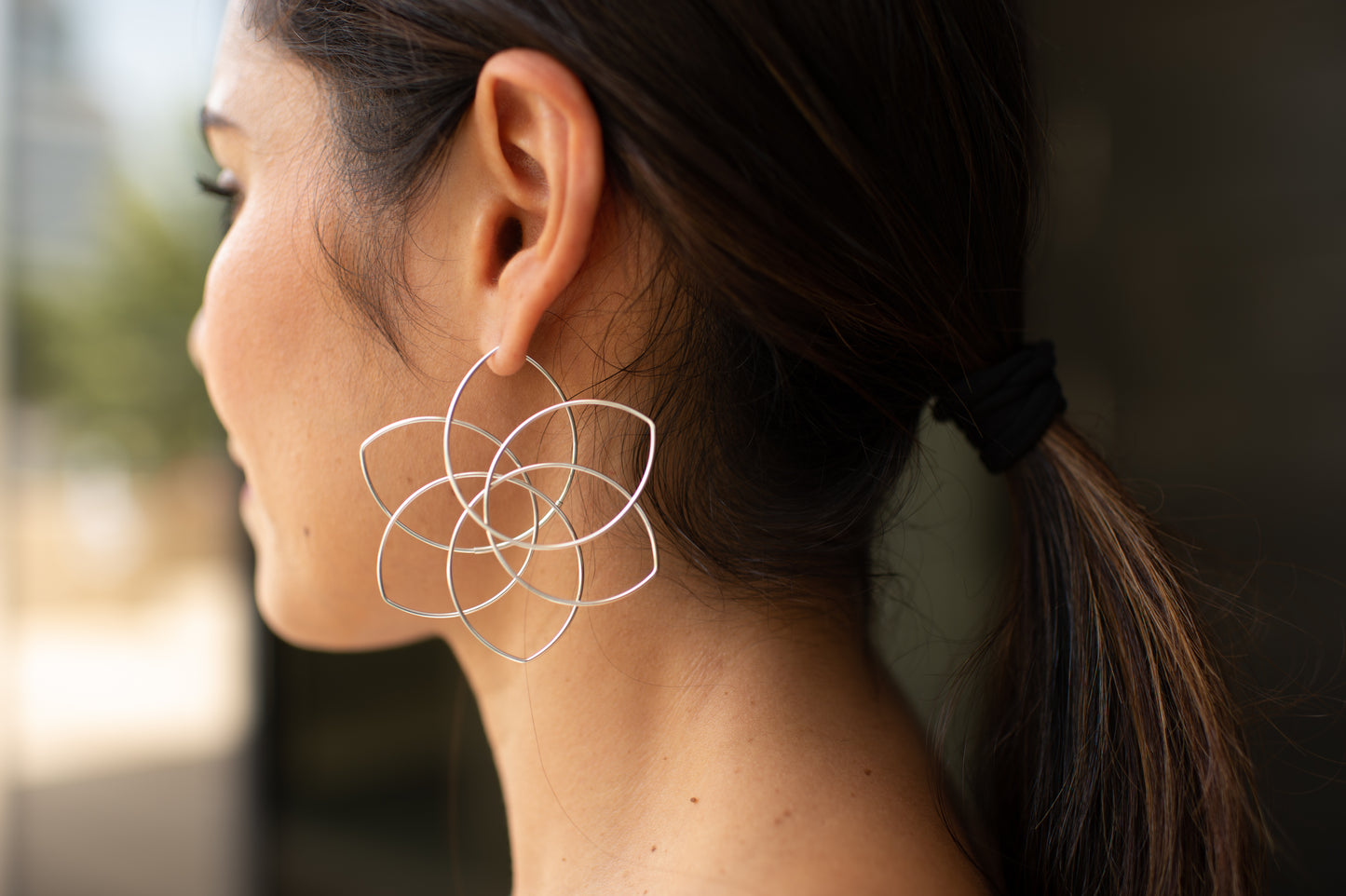A model with long dark silky hair shows off a pair of flower earrings by American-Belgian jewelry designer Hannah Tomoko. Like a geometric 6-petal flower, these earrings are balanced not only visually but also when they are worn: they are large but light and they close in a nearly invisible tubing to enhance the continuity of the design. Each earring is made of one continuous sterling silver wire that is meticulously curved and bent.